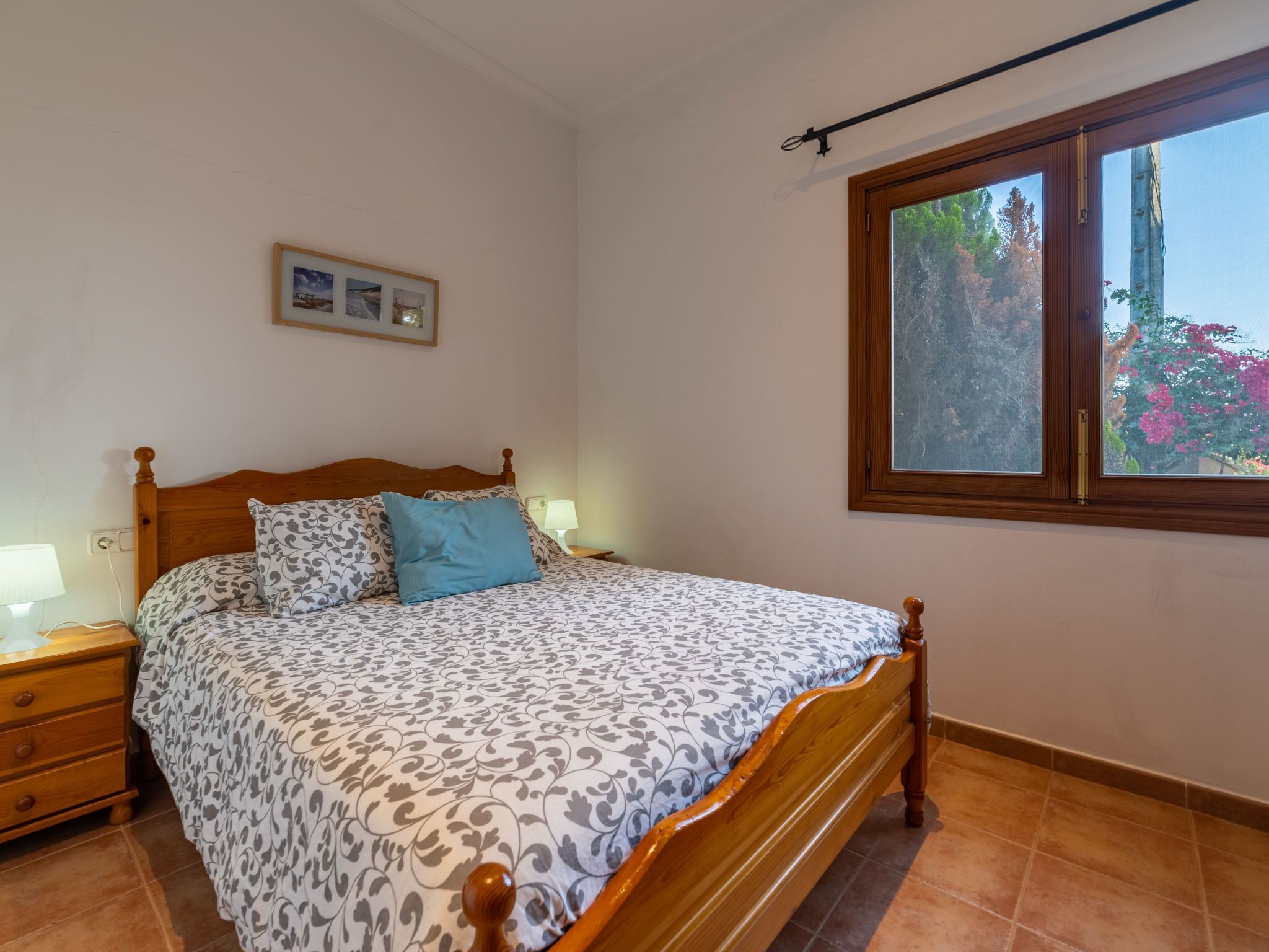 Une chambre à coucher avec un lit en bois, une table de chevet, une lampe et une fenêtre donnant sur des arbres et des fleurs.