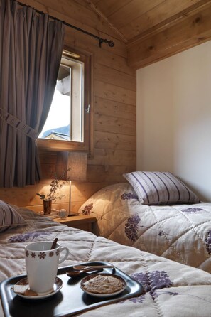 A cozy bedroom with wooden walls, a window with curtains, a bedside table with a lamp, and a tray with a cup and cookies.