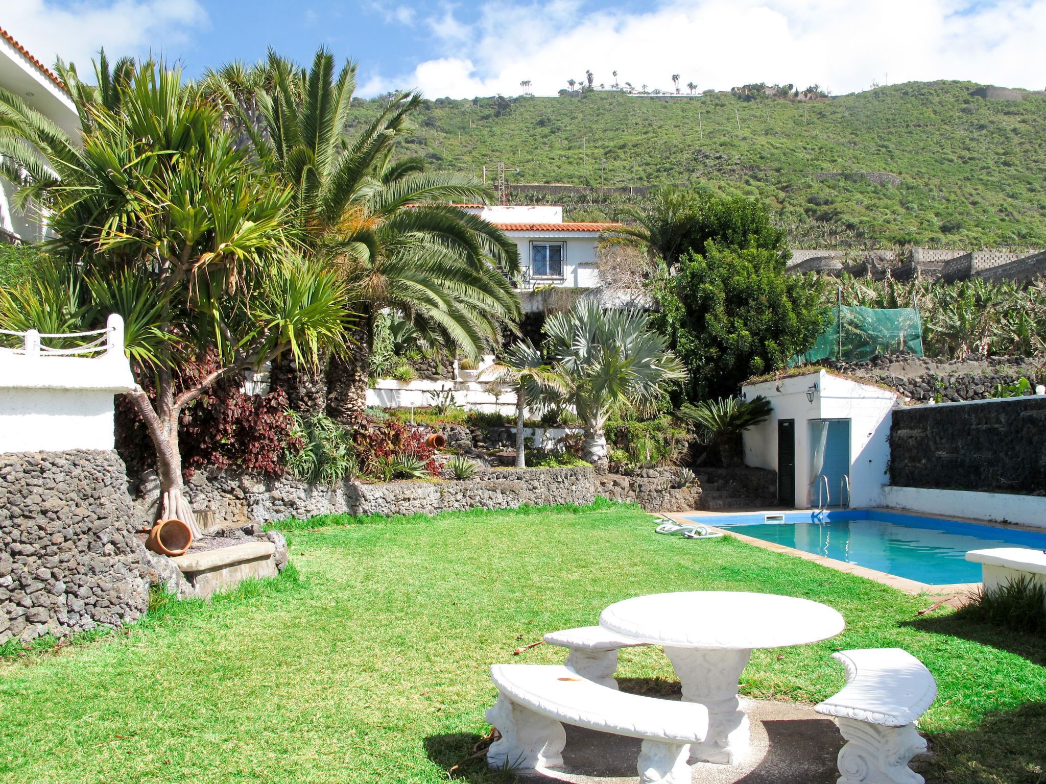 Zona de piscina amb un mur de pedra, un edifici blanc i un jardí amb gespa.