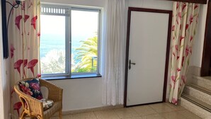 Habitació amb una cadira de vusta, una finestra que dona a l'oceà i una porta amb cortines florals.