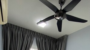 Ein ordentlich bezogenes Bett mit einem weißen Kopfkissen und einem zusammengerollten Handtuch, ein Deckenventilator und ein Fenster mit durchsichtigen Vorhängen.