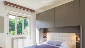 Une chambre moderne avec un grand lit, des rangements intégrés, un radiateur et une fenêtre donnant sur un espace verdoyant.