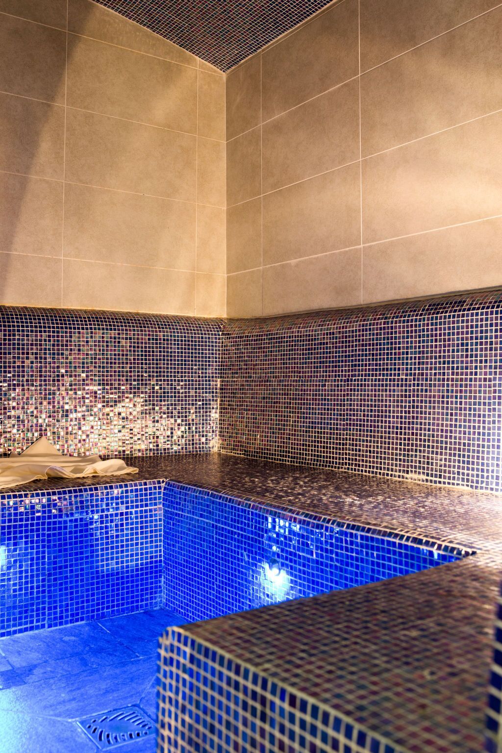 Een wellnessruimte met een blauw betegeld zwembad, beige wanden en een mozaïekpatroon.