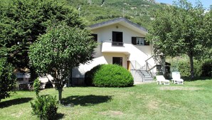 Uma casa de dois andares com varanda, cercada por vegetação e montanhas ao fundo.