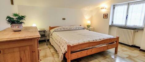 A bedroom with a wooden bed, a bedside table, a lamp, a radiator, and a window with curtains.