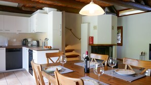 Un comedor con una mesa de madera dispuesta para cuatro personas, rodeada de sillas. Se ve una cocina al fondo, con armarios blancos y un salpicadero de azulejos.