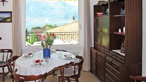 A dining area with a table set for four, a view of a lake with mountains, and a wooden cabinet with decorative items. - Elisabetta (GLA116) by Interhome (Gera Lario)