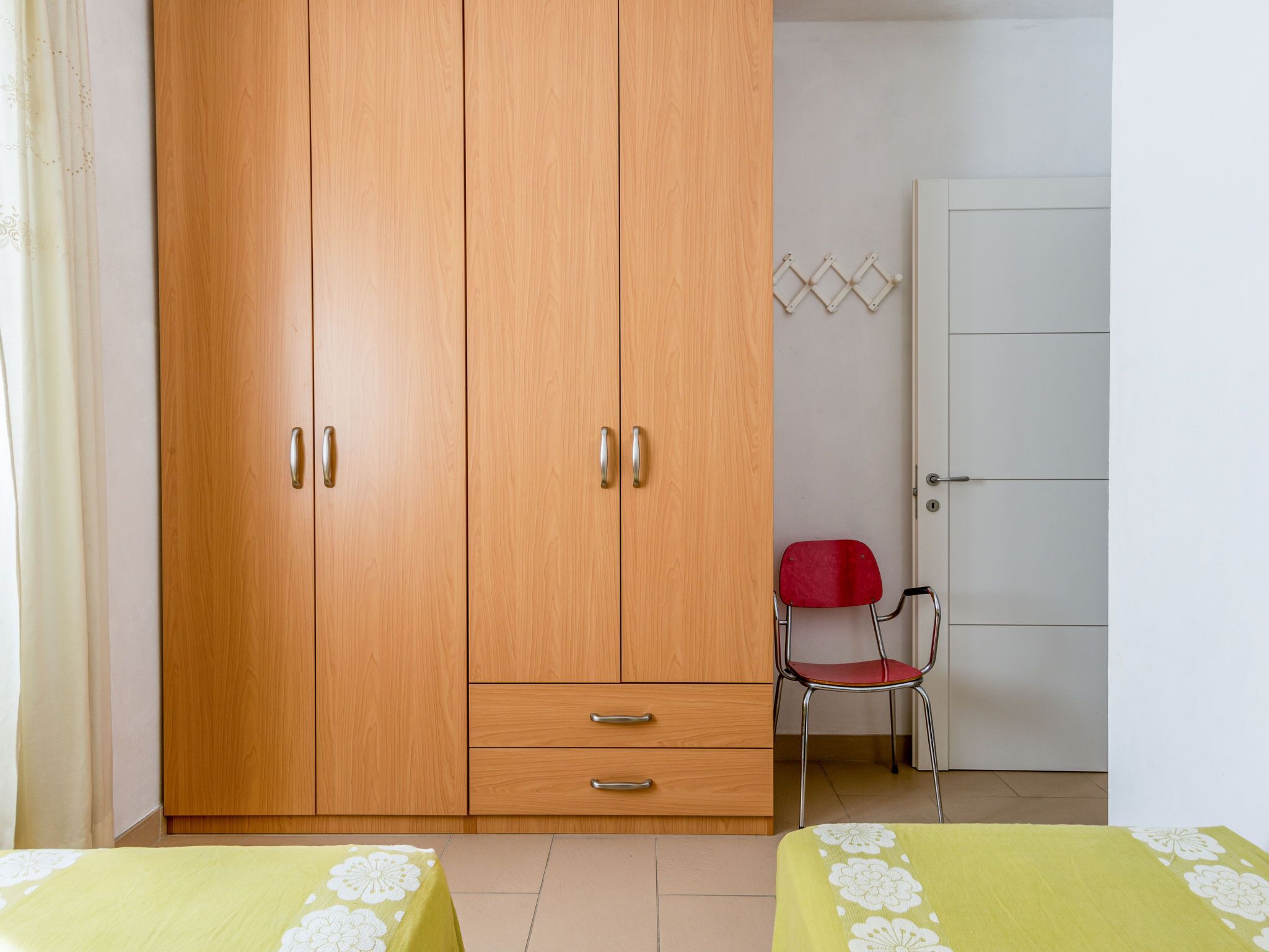 A room with a wooden wardrobe, a red chair, and two beds with floral bedspreads.