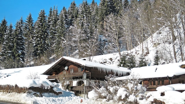 A wooden chalet with a snow-covered roof, surrounded by a forest and a snow-covered hill.