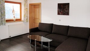 Una sala de estar con un sofá marrón oscuro, una mesa de centro de vidrio, una puerta de madera y una ventana con cortinas.