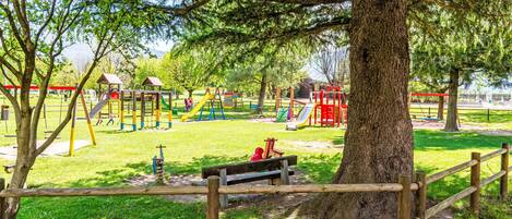 Un parque infantil con diversas estructuras de juego, un banco y un árbol grande al frente.