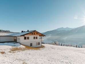 Un edificio de dos pisos con techo inclinado, situado en una ladera nevada con vistas a montañas al fondo.