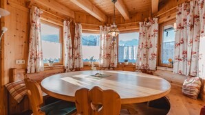 A wooden interior with a round table and chairs, large windows with floral curtains, and a view of mountains. - Geislhof by Interhome (Bramberg am Wildkogel)