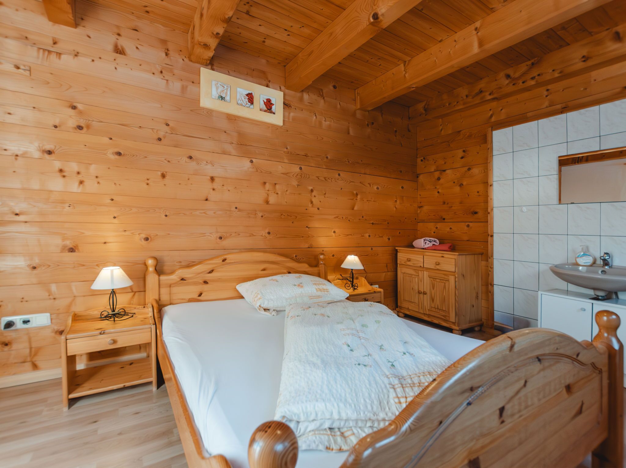 A wooden cabin room with a bed, two bedside tables with lamps, a wooden cabinet, and a bathroom with a sink and mirror.