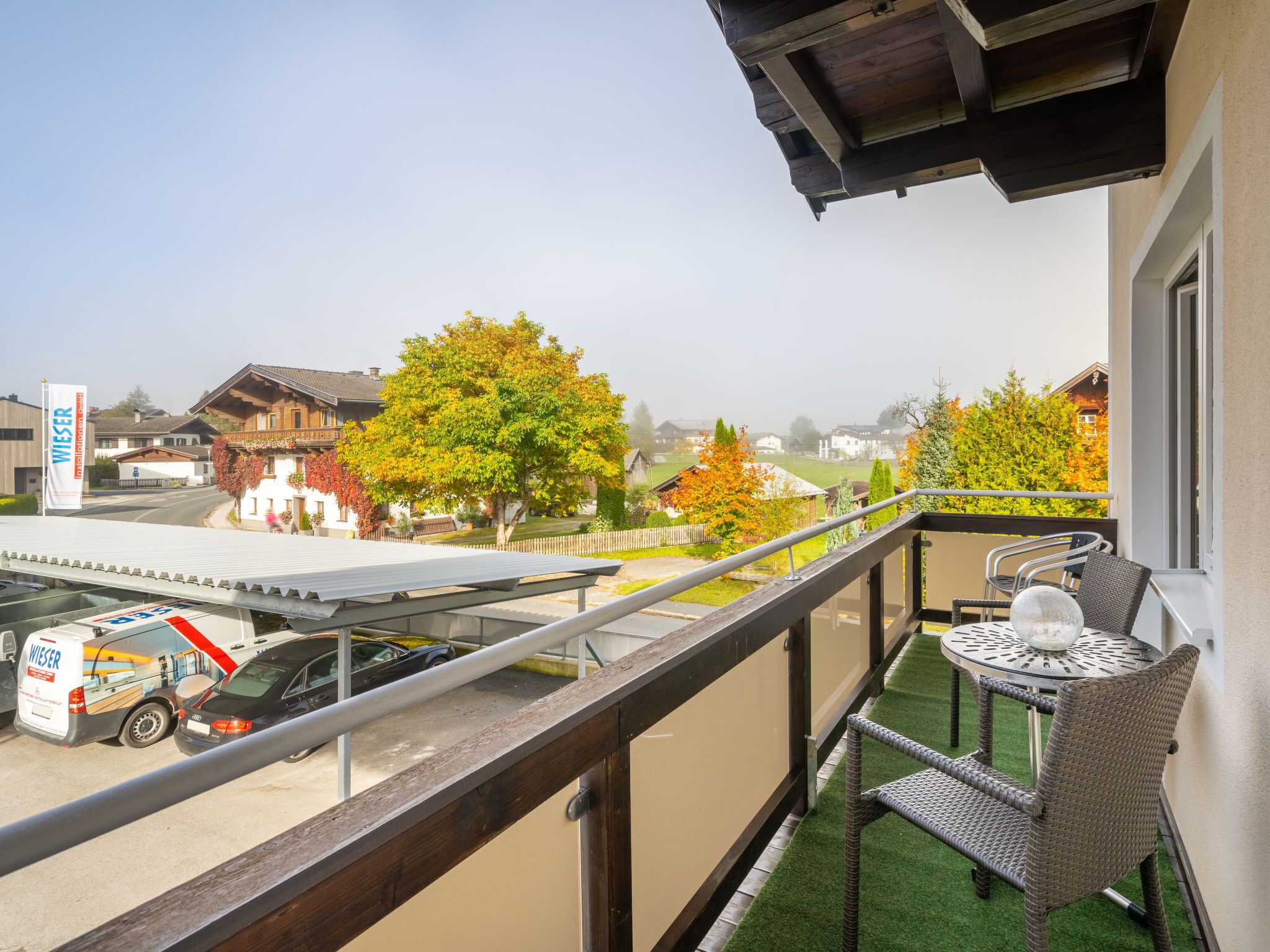 A balcony with a view of a parking area, a building with a wooden facade, and a tree with autumn foliage.