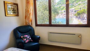 A room with a large window offering a view of a hillside village, a black leather chair with a floral pillow, and a radiator on the wall. - Apartment Betti by Interhome (Domaso)