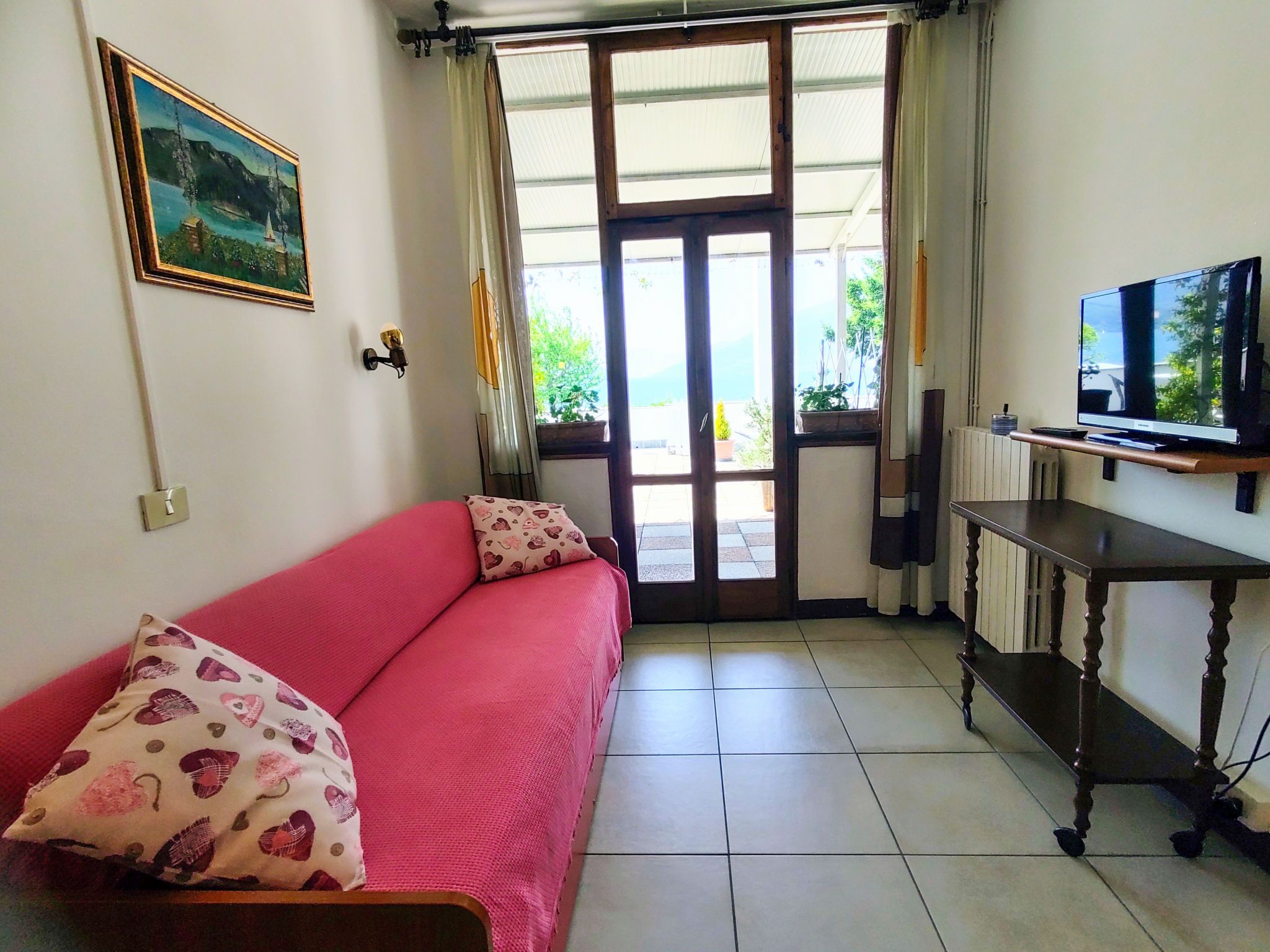 Quarto com sofá vermelho, televisão sobre um suporte de madeira e porta que dá para o exterior.