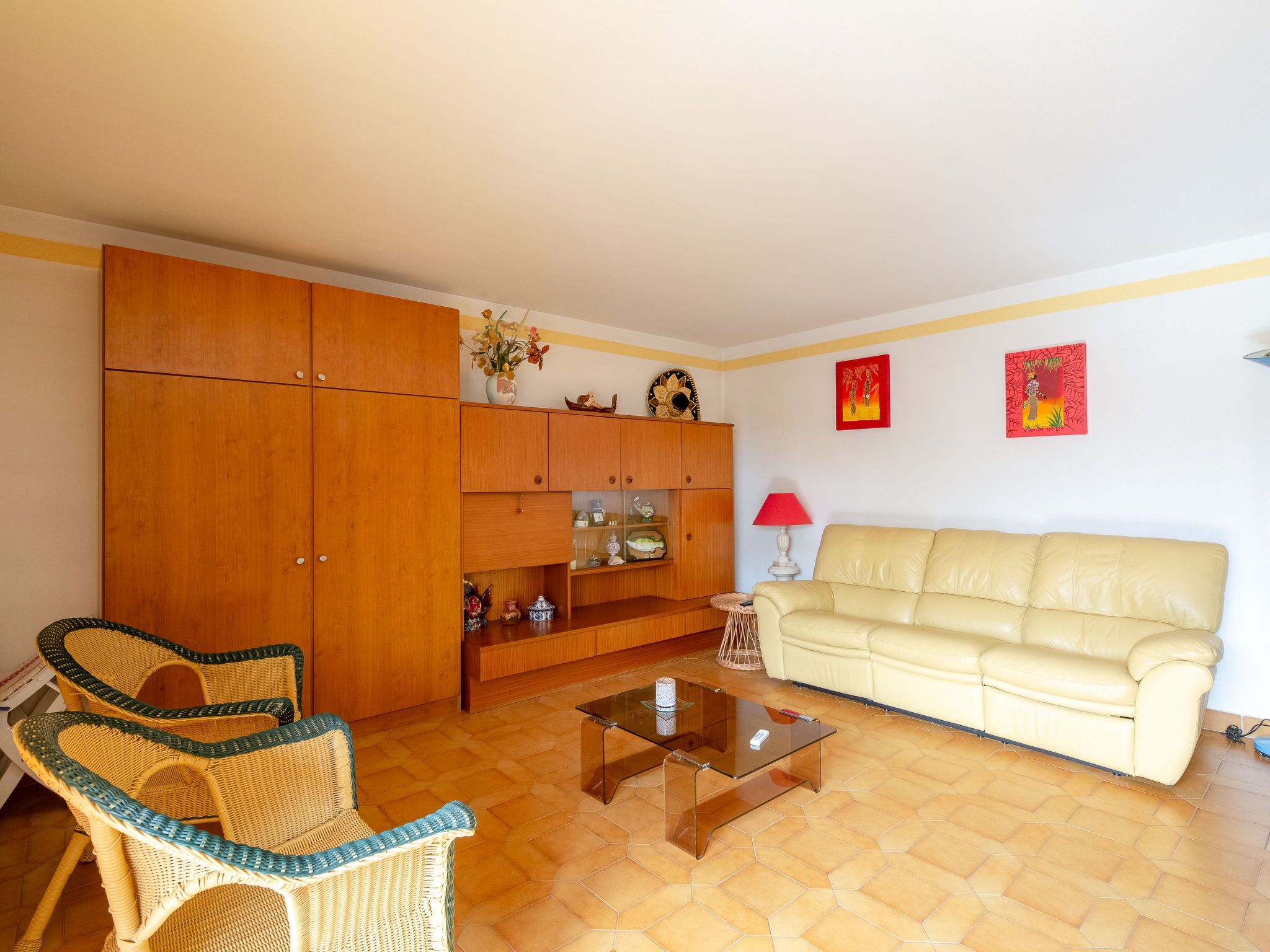 A living room with a beige sofa, wicker chairs, a glass coffee table, wooden cabinets, and two red wall art pieces.