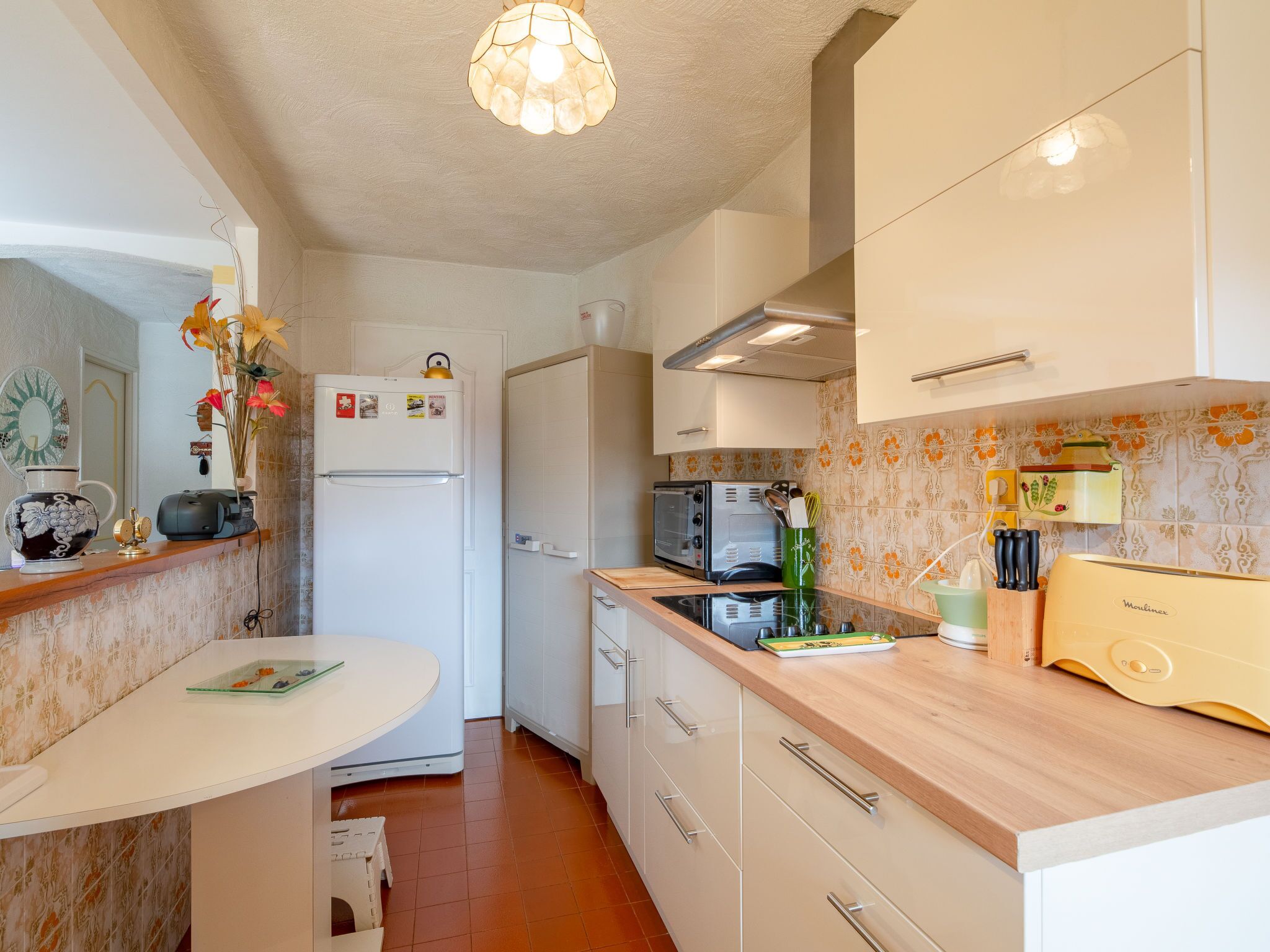 A compact kitchen with a white refrigerator, a microwave, and a toaster on the countertop. There is a small dining table with a vase of flowers on the left.