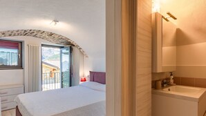 A bedroom with a bed, a nightstand, a window with curtains, a balcony, and a bathroom with a sink and mirror. - Marcello by Interhome (Gravedona)