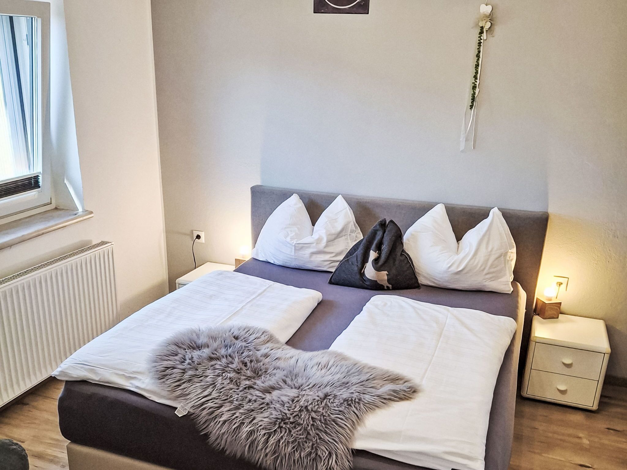 Un letto rifatto con lenzuola bianche, una coperta in pelliccia grigia e un mix di cuscini bianchi e neri. C'è un comodino con una lampada e una piccola pianta.