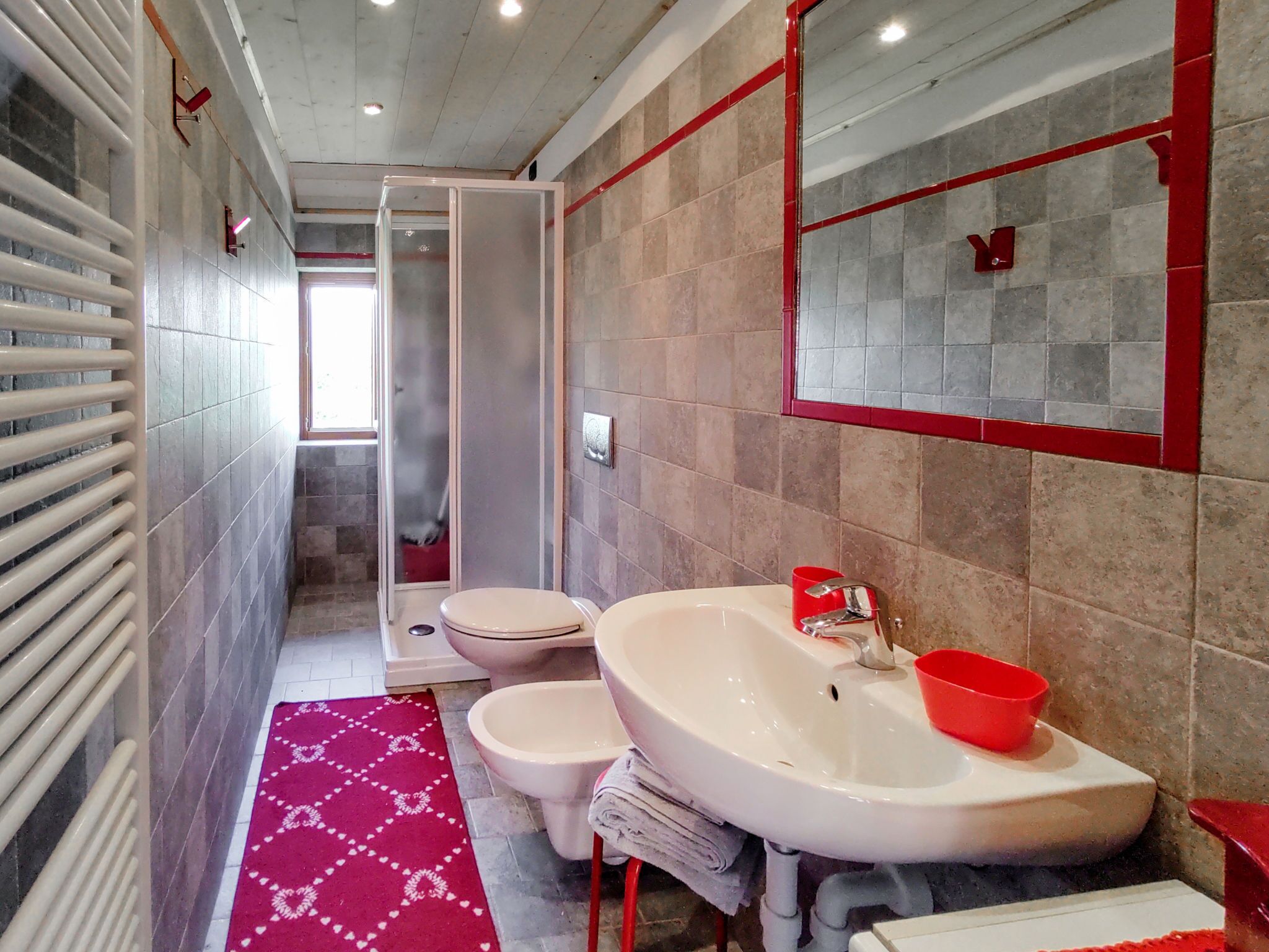 Un baño moderno con un lavamanos blanco, un lavabo rojo y un cubo rojo. Hay un inodoro blanco y un box de ducha con puerta de vidrio. Una alfombra estampada en rojo y blanco está en el suelo.