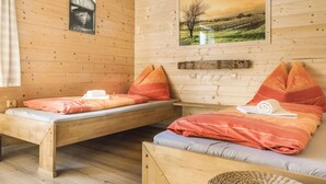 Ein Zimmer in einer Holzhütte mit zwei Betten, jeweils mit orange-weißer Bettwäsche, einem gerahmten Bild eines Weinbergs und einem kleinen Fenster.