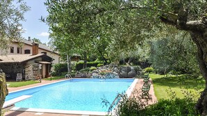 A rectangular swimming pool with a tiled deck, surrounded by lush greenery and a stone building with a tiled roof.