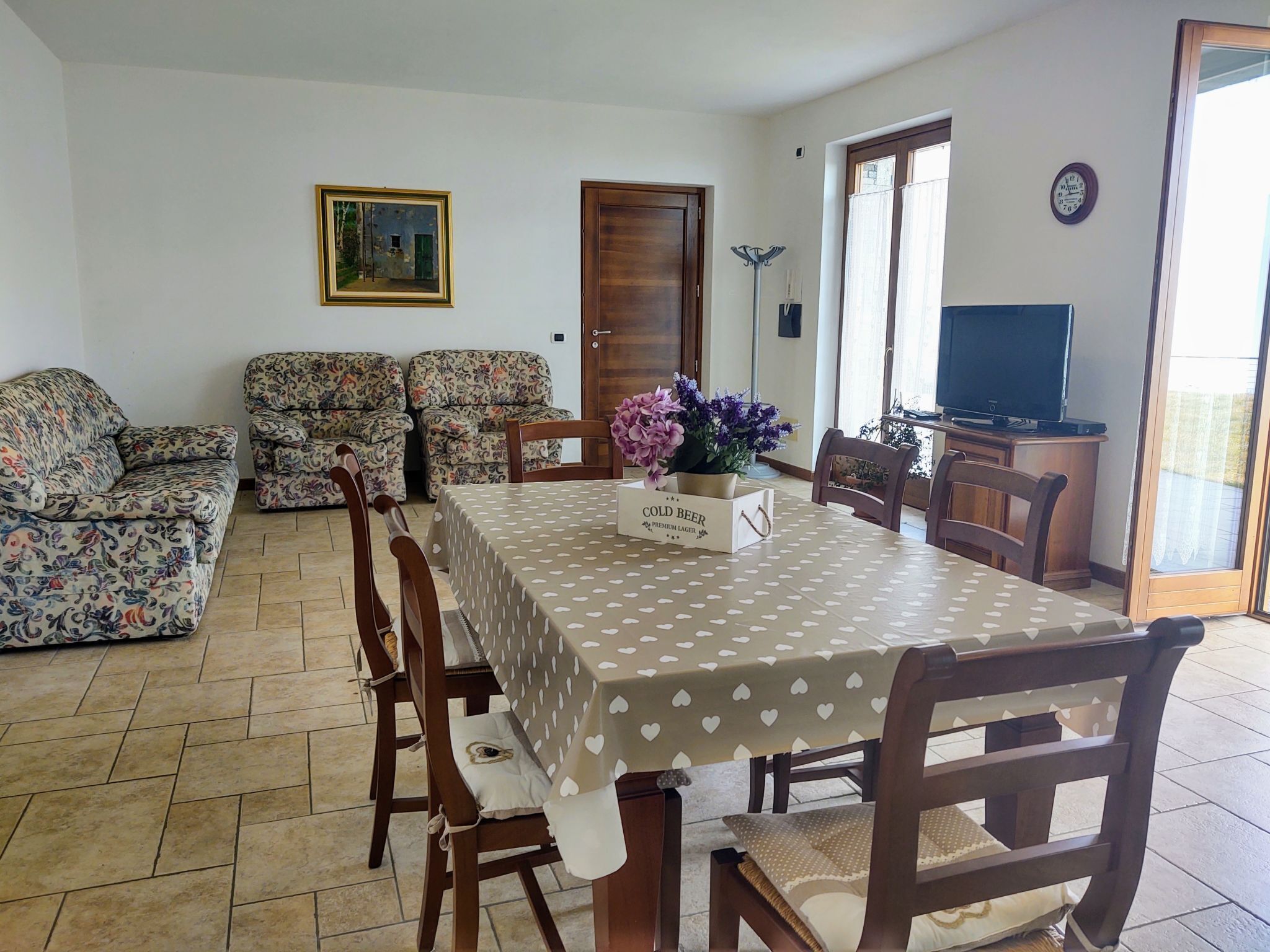 Uma área de refeições com uma mesa posta para quatro pessoas, cercada por sofás com estampa floral. Uma televisão está visível ao fundo.