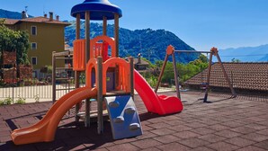 A playground with slides and climbing structures, surrounded by buildings and a mountainous backdrop.