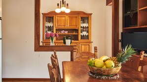 A dining area with a wooden table, chairs, a fruit bowl, and a kitchen in the background.