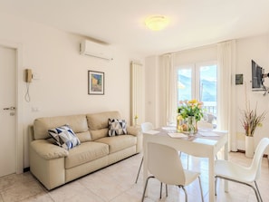 A living room with a beige sofa, a dining table with chairs, a window with curtains, and a wall-mounted air conditioner.