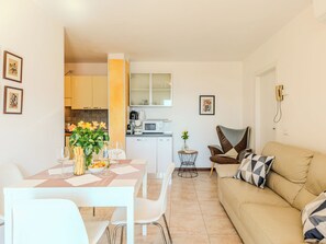 A modern living room with a beige sofa, a dining table with chairs, a kitchen area with cabinets and appliances, and artwork on the walls.