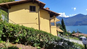 A yellow building with a balcony, surrounded by greenery and overlooking a lake with mountains in the background.