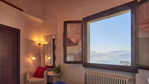 A hotel room with a bed, a chair, a small table with a plant, a lamp, and a window with a view of the sea and mountains. - Bellavista by Interhome (Gravedona)