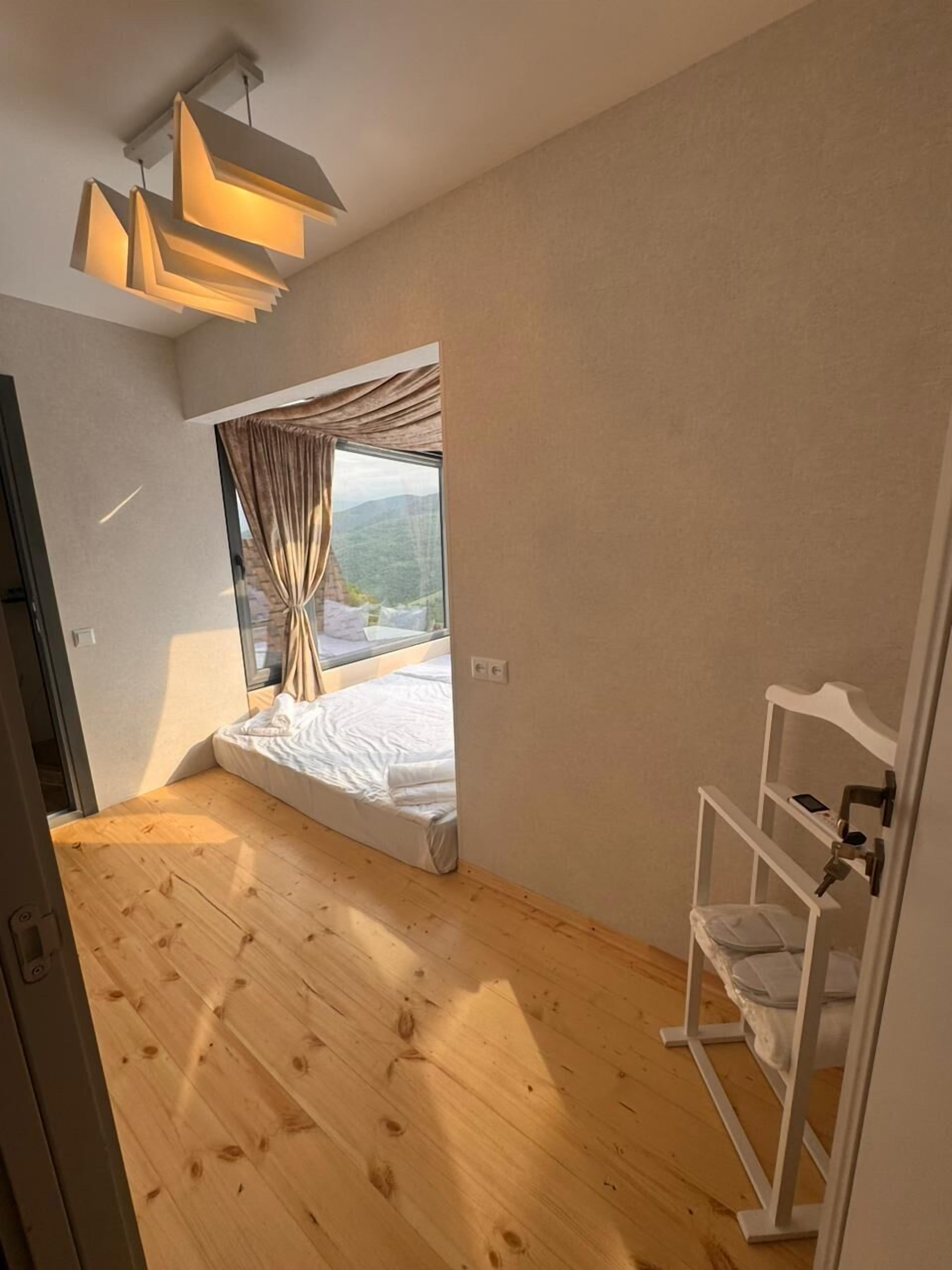 A bedroom with a wooden floor, a bed with white bedding, a window with curtains, and a white shelf unit.