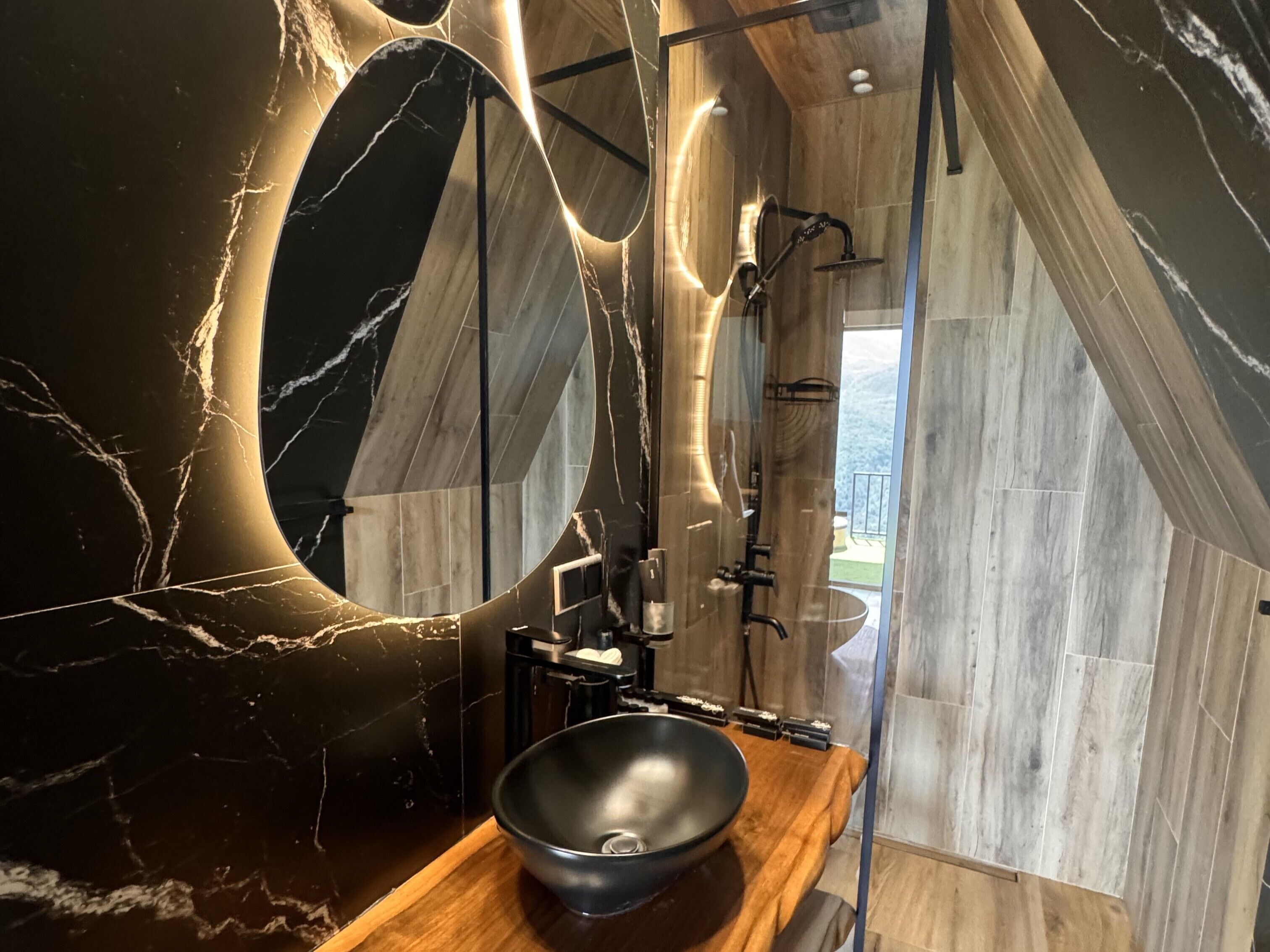 Un baño moderno con un espejo grande, un lavabo negro y suelo de madera.