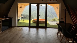 A room with a wooden floor, a dining table with chairs, a small kitchen area, and a large sliding glass door leading to an outdoor terrace with a view of a mountain. - Lost in Sky (Yenokavan)