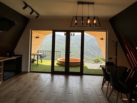 Una habitación con piso de madera, una mesa de comedor con sillas, una pequeña cocina y un amplio ventanal corredero que da a una terraza exterior con vistas a la montaña.