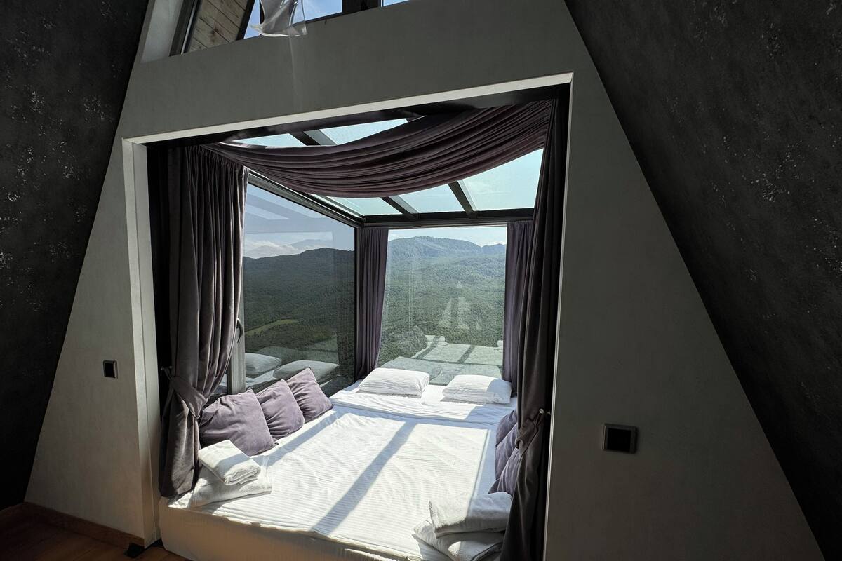 A bedroom with a bed, curtains, and a view of a mountainous landscape through a large window.