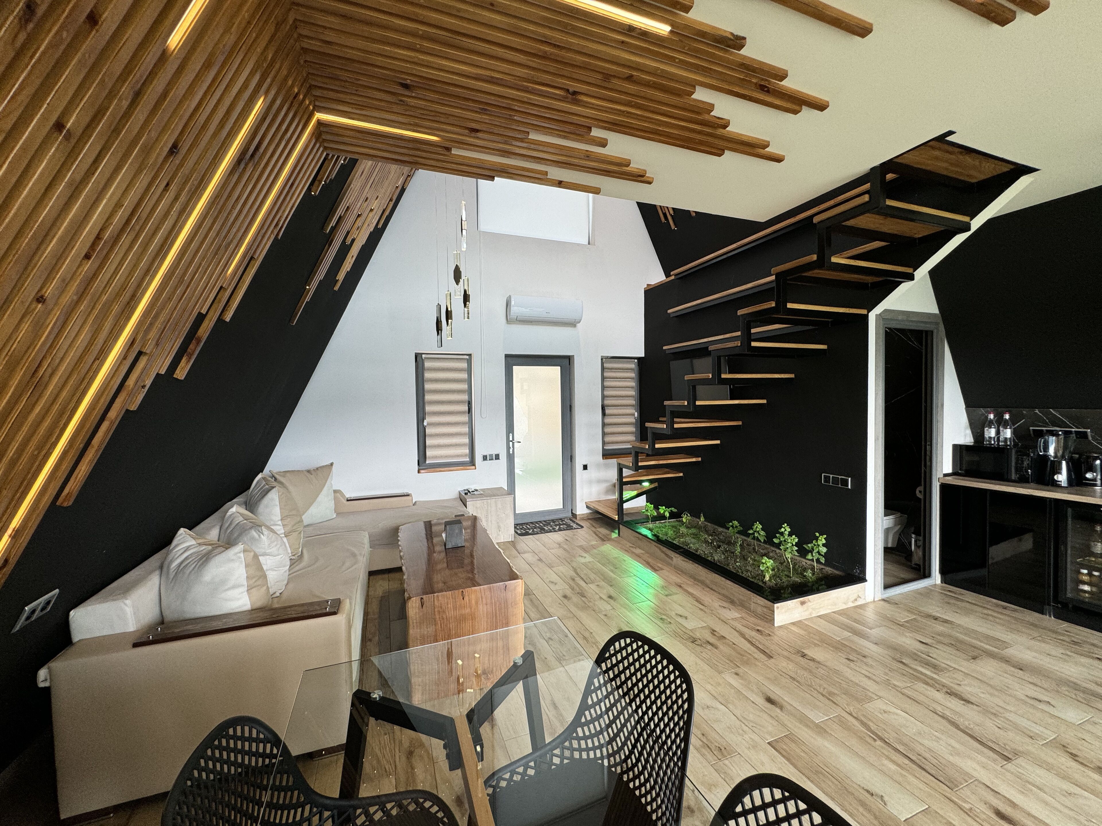 A modern loft-style living space with a sloped ceiling, a glass dining table, a sofa, and a kitchen area with a built-in stove.