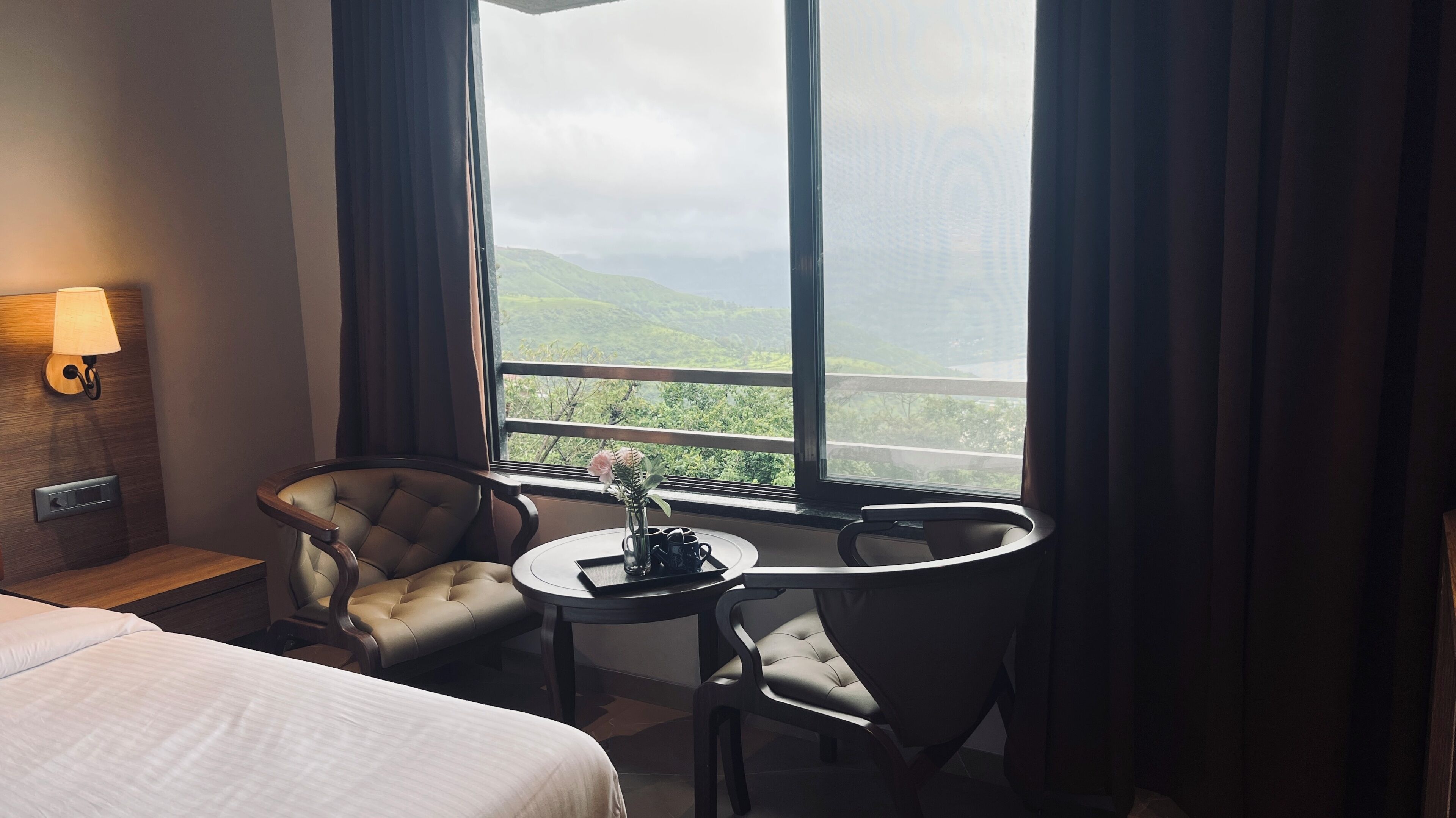 A hotel room with a bed, a seating area with two chairs, a small table with a vase and cups, and a window with a view of greenery.