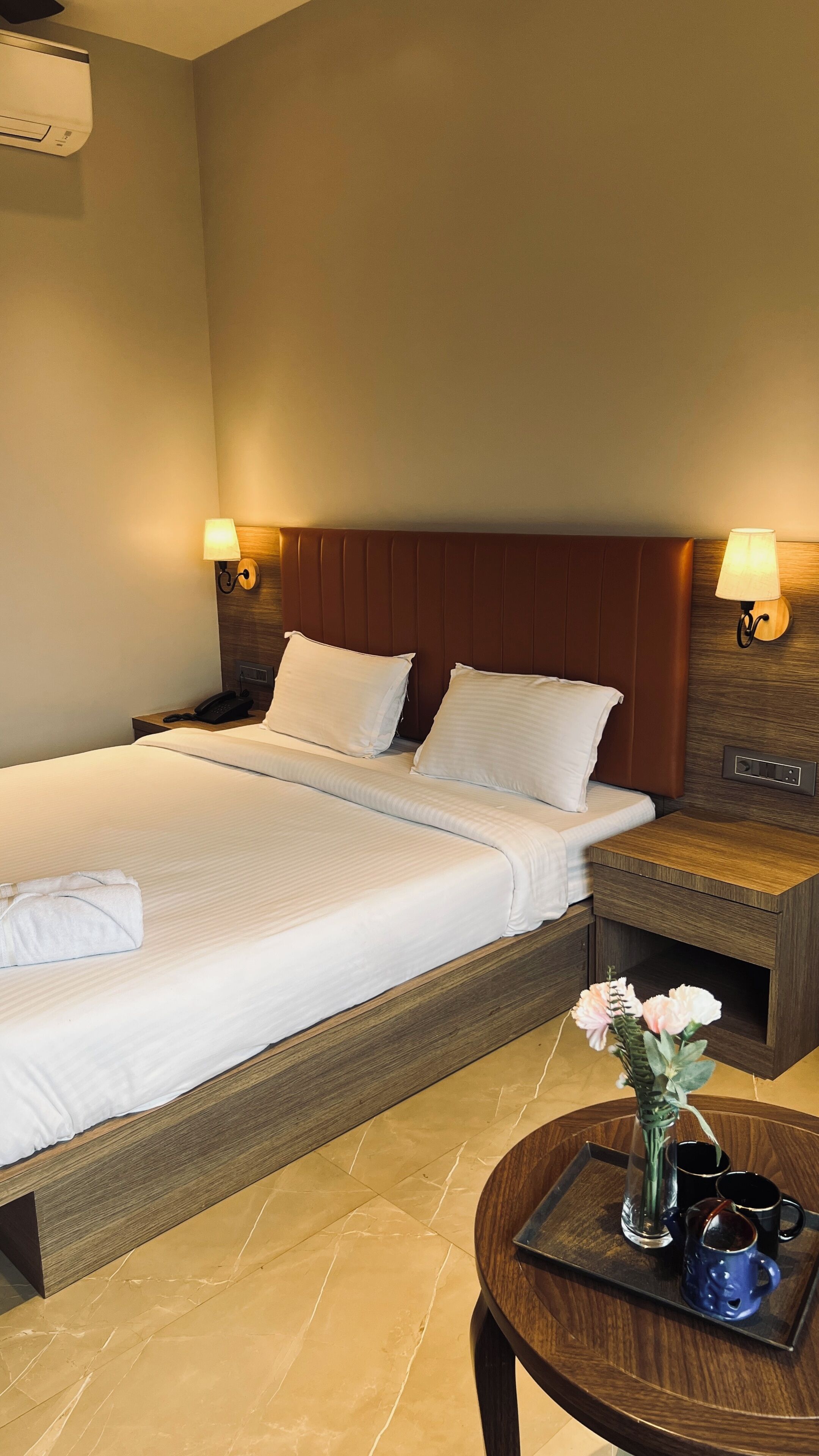 Ein Hotelzimmer mit einem großen Bett, Nachttischlampen, einem Telefon am Nachttisch, einem kleinen Tisch mit einer Vase und Tassen sowie einer Wand-Klimaanlage.