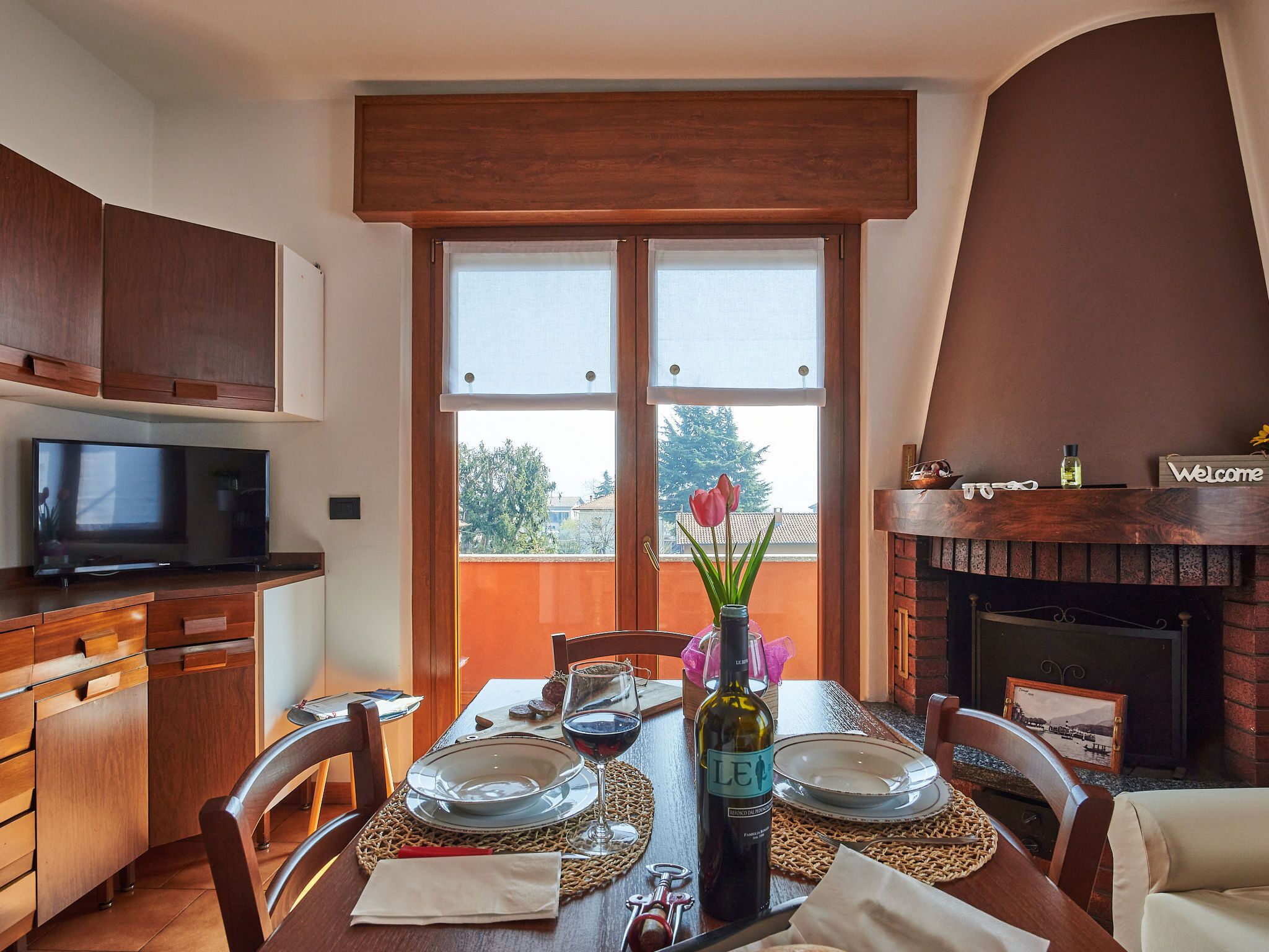 A dining area with a wooden table set for two, a television on a wooden cabinet, and a fireplace with a 'Welcome' sign.