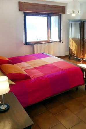 A bed with a colorful quilt, a bedside table with a lamp, a radiator, and a cabinet with a mirror. - Apartment Nicole by Interhome (Porlezza)