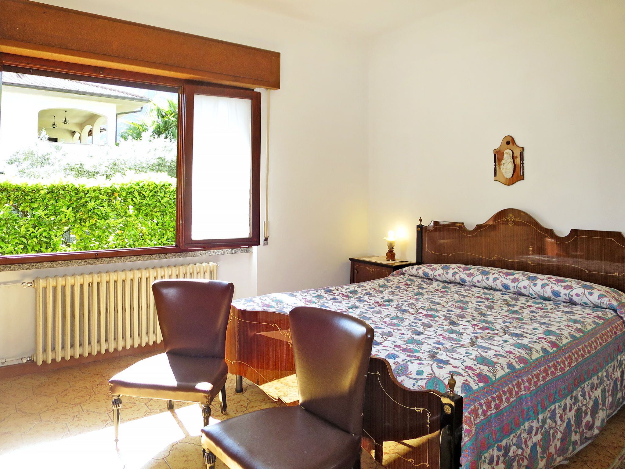 A bedroom with a bed, two chairs, a nightstand, a lamp, a window with a view of greenery, and a radiator.