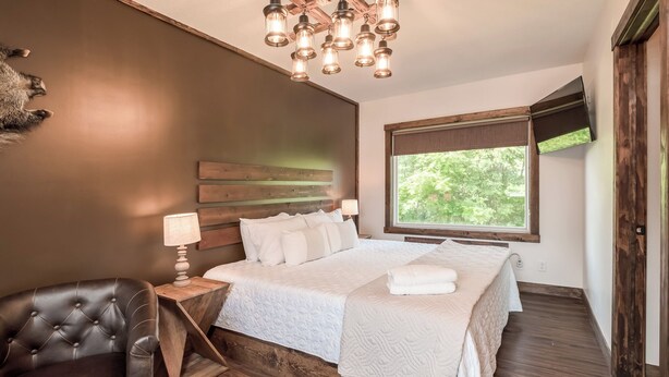Une chambre avec un grand lit, une tête de lit en bois, un fauteuil en cuir, une table de chevet en bois, une lampe sur pied et une fenêtre donnant sur un paysage verdoyant.