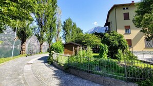 A cobblestone path leading to a wooden cabin with a metal fence, surrounded by lush greenery and a mountain in the background. - Punto Lago by Interhome (Lago di Mezzola)