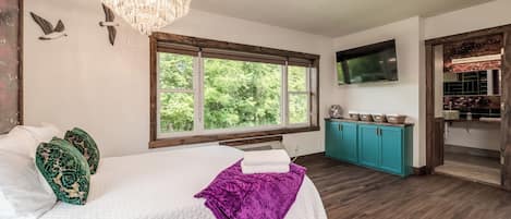 A bedroom with a bed, a chandelier, a window with a view of trees, a television, and a bathroom with a sink and cabinets.