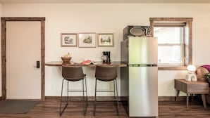A compact kitchen with a refrigerator, two bar stools, a coffee maker, and framed pictures on the wall.